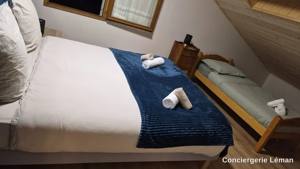 Chambre mansardée : lit double fait avec plaid bleu nuit, serviettes roulées, lit d’appoint préparé
